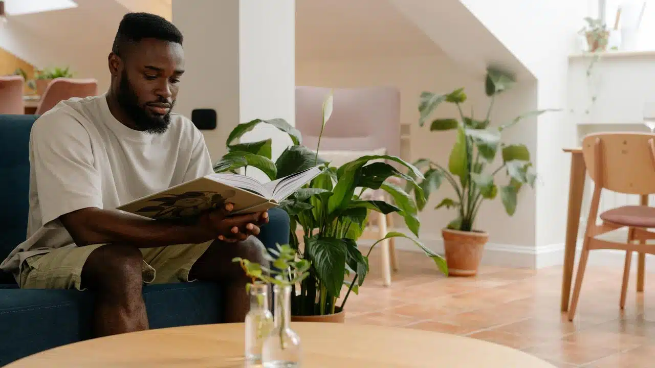 A man sitting on a couch reading a book in a bright room with plants.