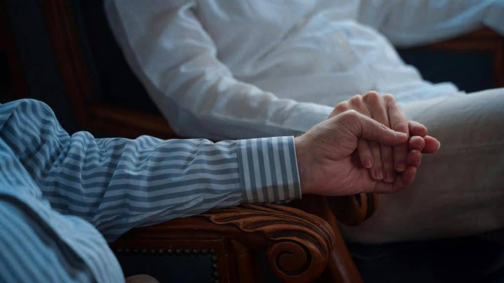 A couple sits holding hands on wooden armchairs.