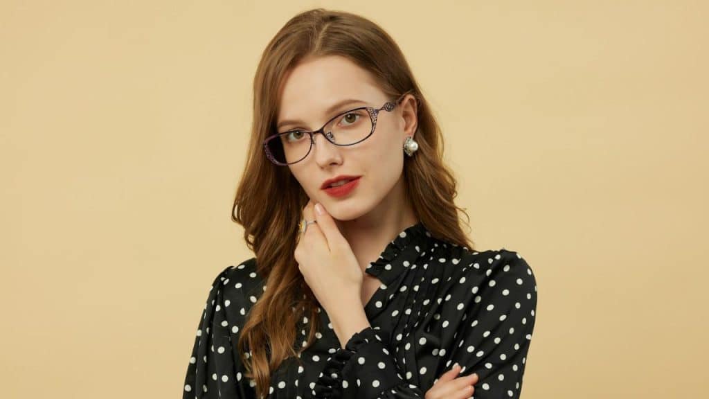 A woman with long brown hair wears glasses and a polka-dot blouse against a plain background.