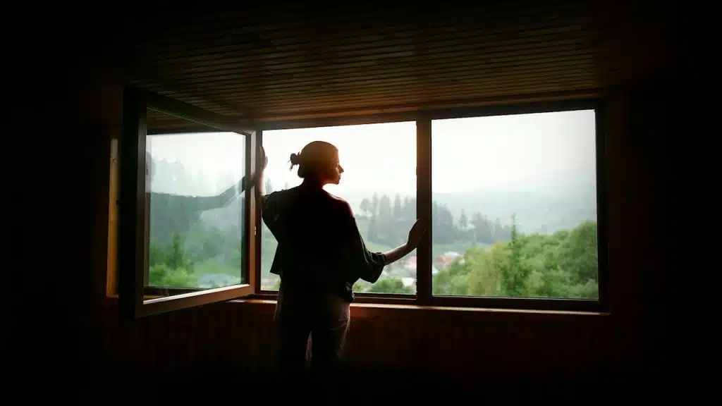 Silhouette of woman in sunshine at big wooden window with view of sunset in mountains.