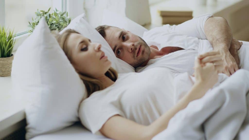 A couple lying in bed; the woman is looking away and the man is looking at her with concern.