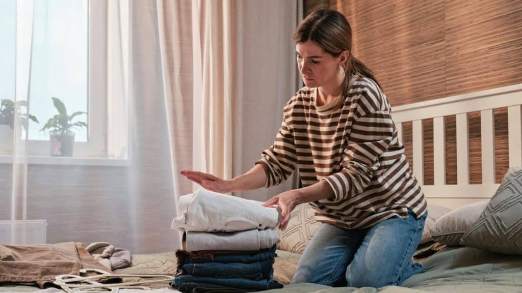 Woman kneeling on a bed, folding and stacking clothes in a bedroom.
