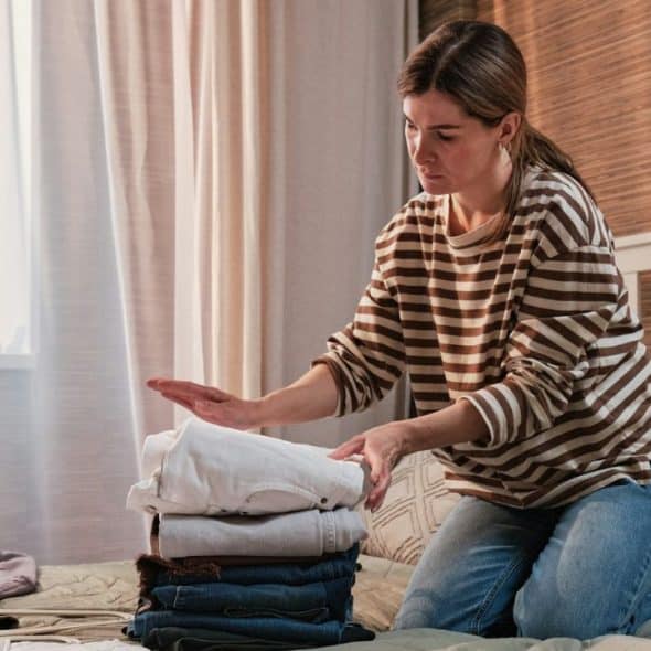 Woman kneeling on a bed, folding and stacking clothes in a bedroom.