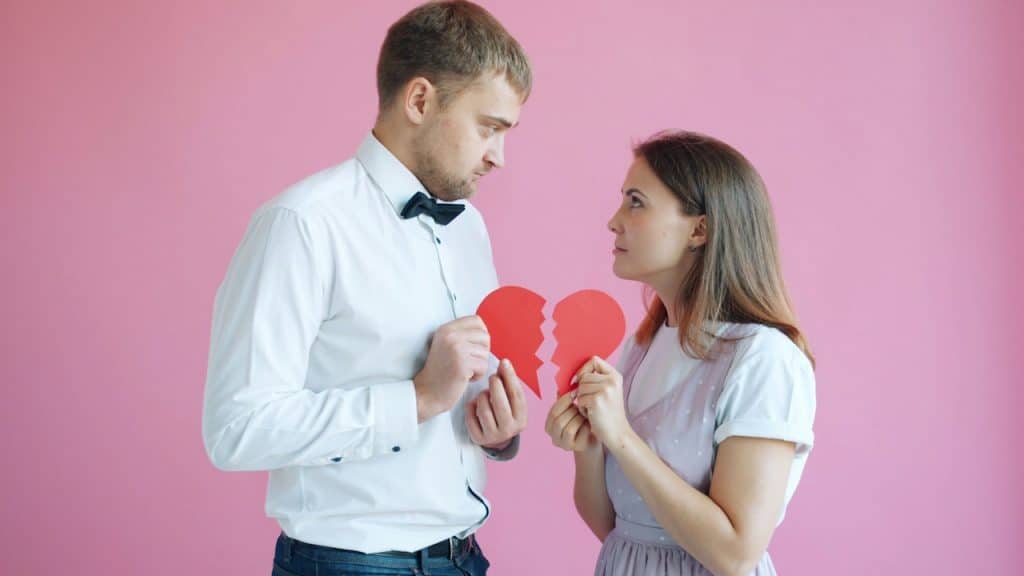 Man and woman facing each other against a pink background, each holding half of a broken red paper heart.
