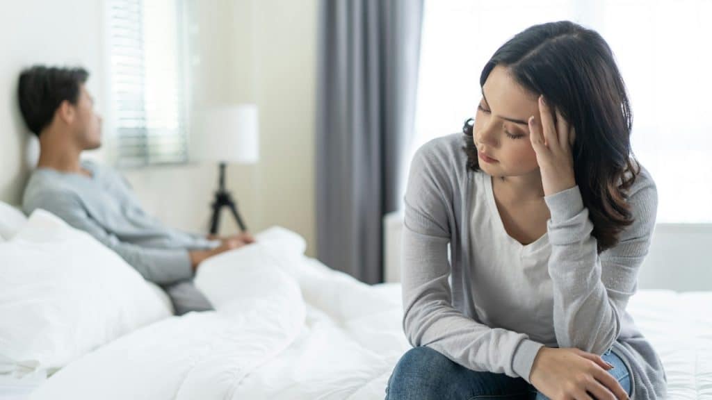 Woman looking upset, sitting on a bed with a man in the background, both facing away from each other.