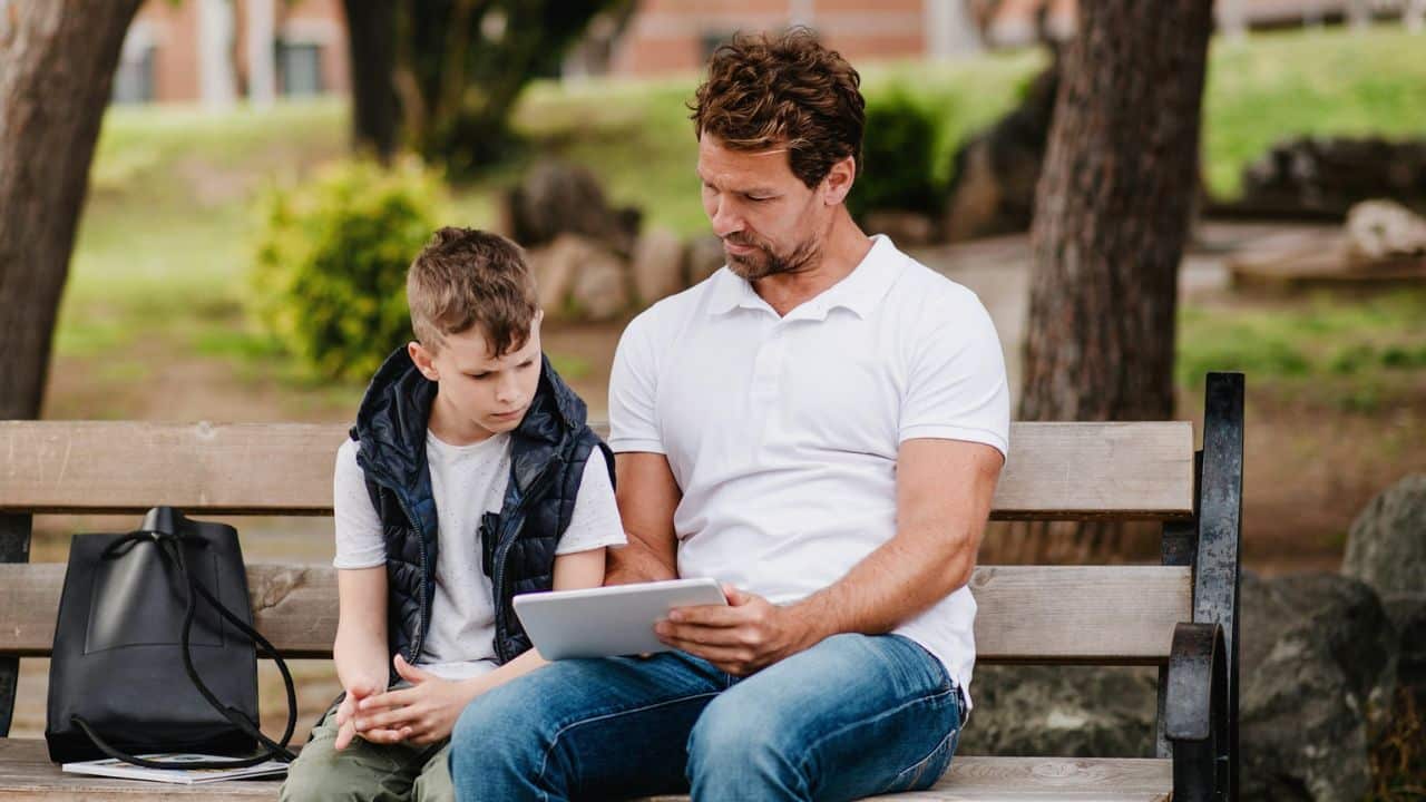 Father and son sitting on a park bench, looking at a tablet together.