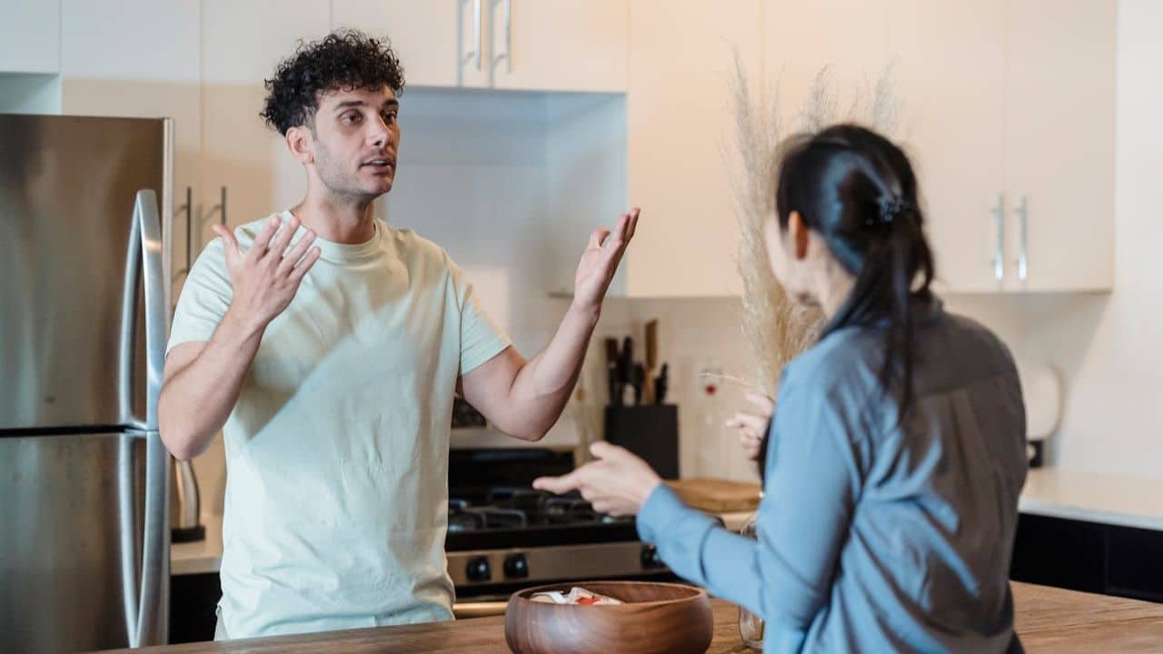 Man with curly hair and a light shirt arguing with a woman in a modern kitchen.