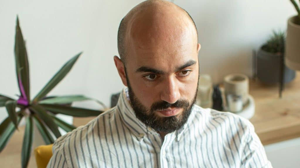 A bald, bearded man in a striped shirt looks down with a serious expression.