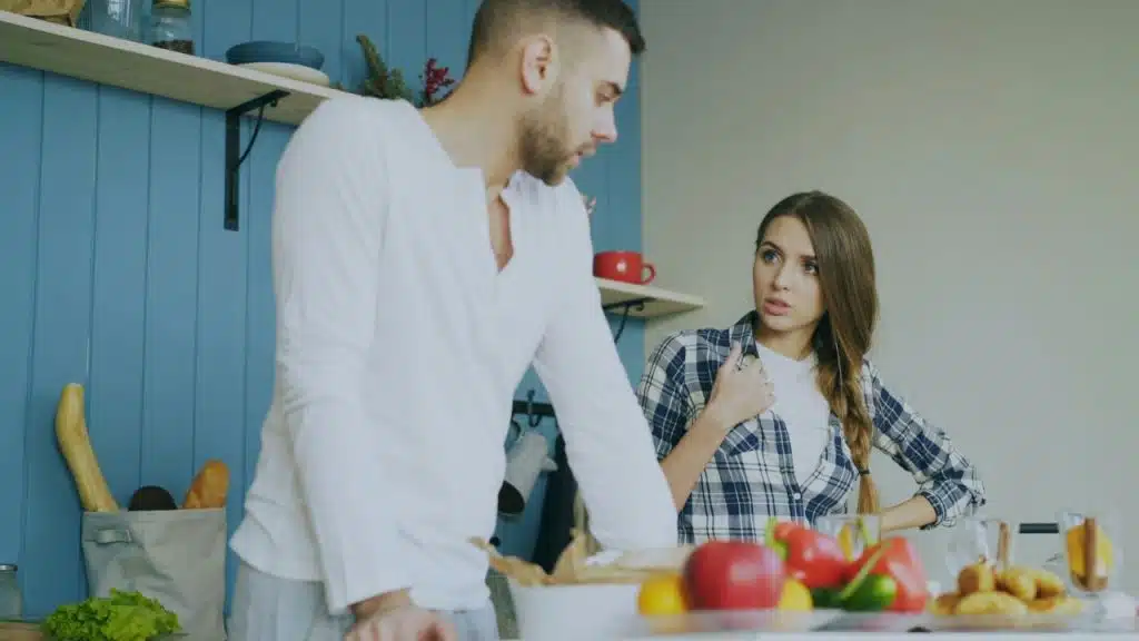 A man and woman are having a serious conversation in a kitchen with food on the counter.