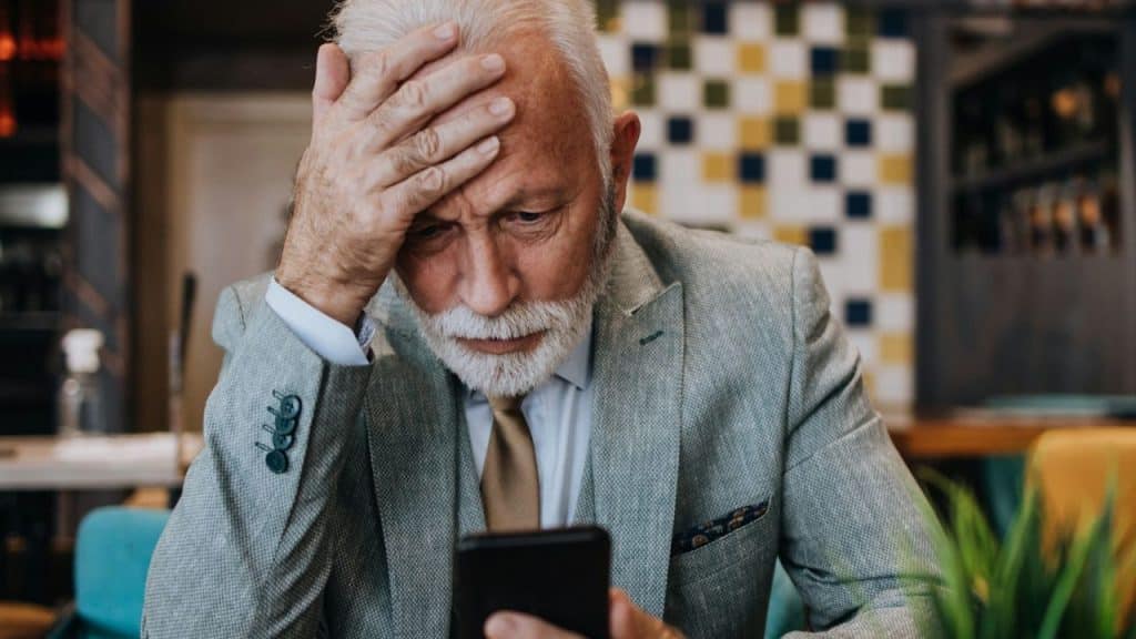 Stressed senior man in a suit and tie holds his forehead while looking at his smartphone.