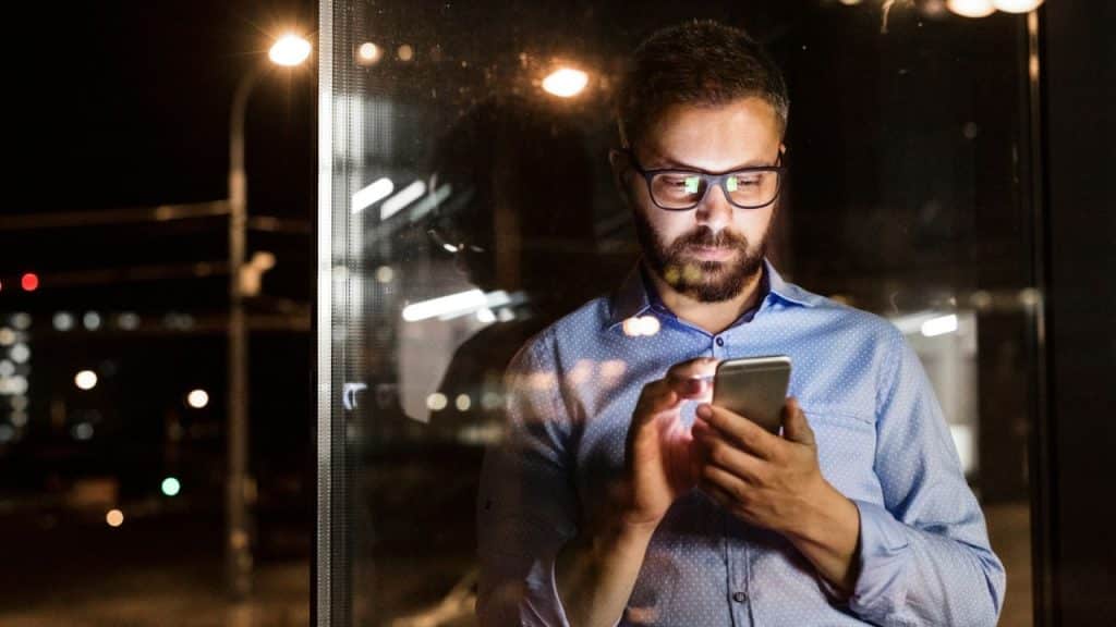Man wearing glasses and a blue shirt looking at a glowing smartphone at night.