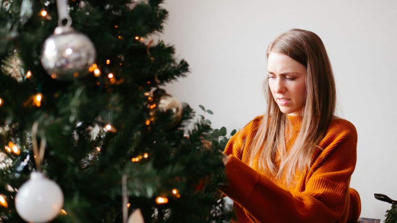 A woman in an orange sweater looks intently while decorating a Christmas tree.