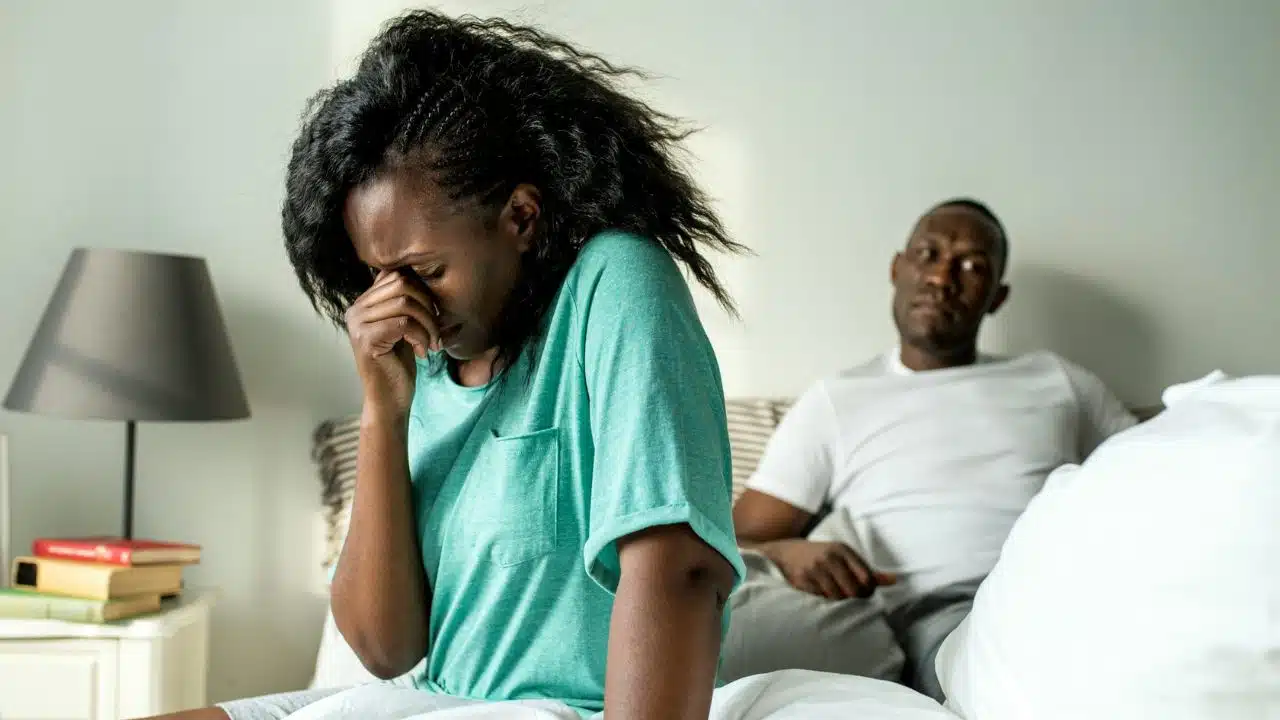 A woman sits on a bed, crying and covering her face, while a man is seated behind her.