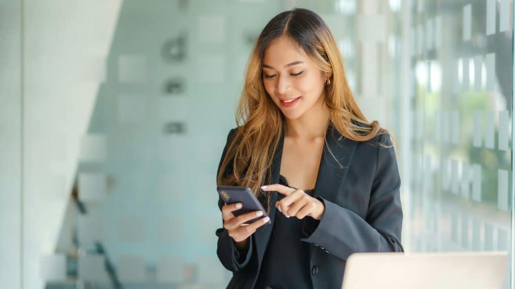A woman in a black blazer smiles while using her smartphone in an office setting.