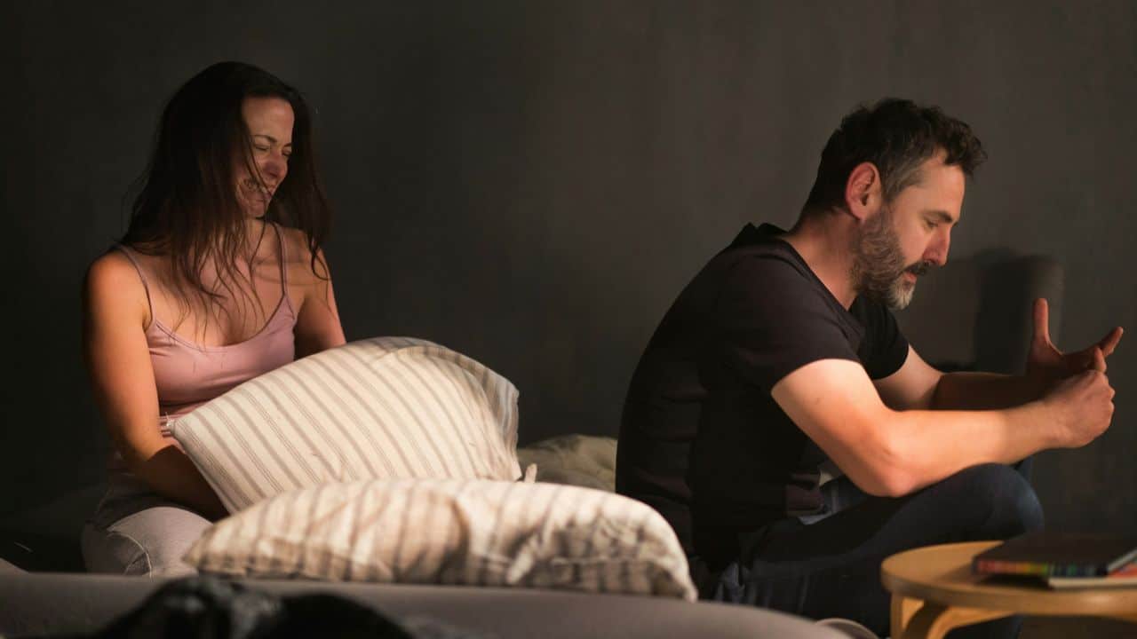 A crying woman holds a pillow next to a man looking down in a darkly lit room.
