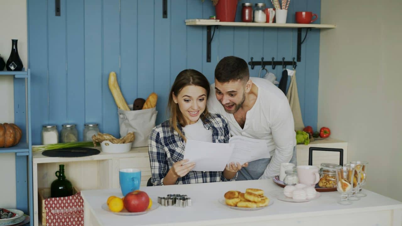 Couple looking excitedly at a paper document in a bright kitchen with blue walls.