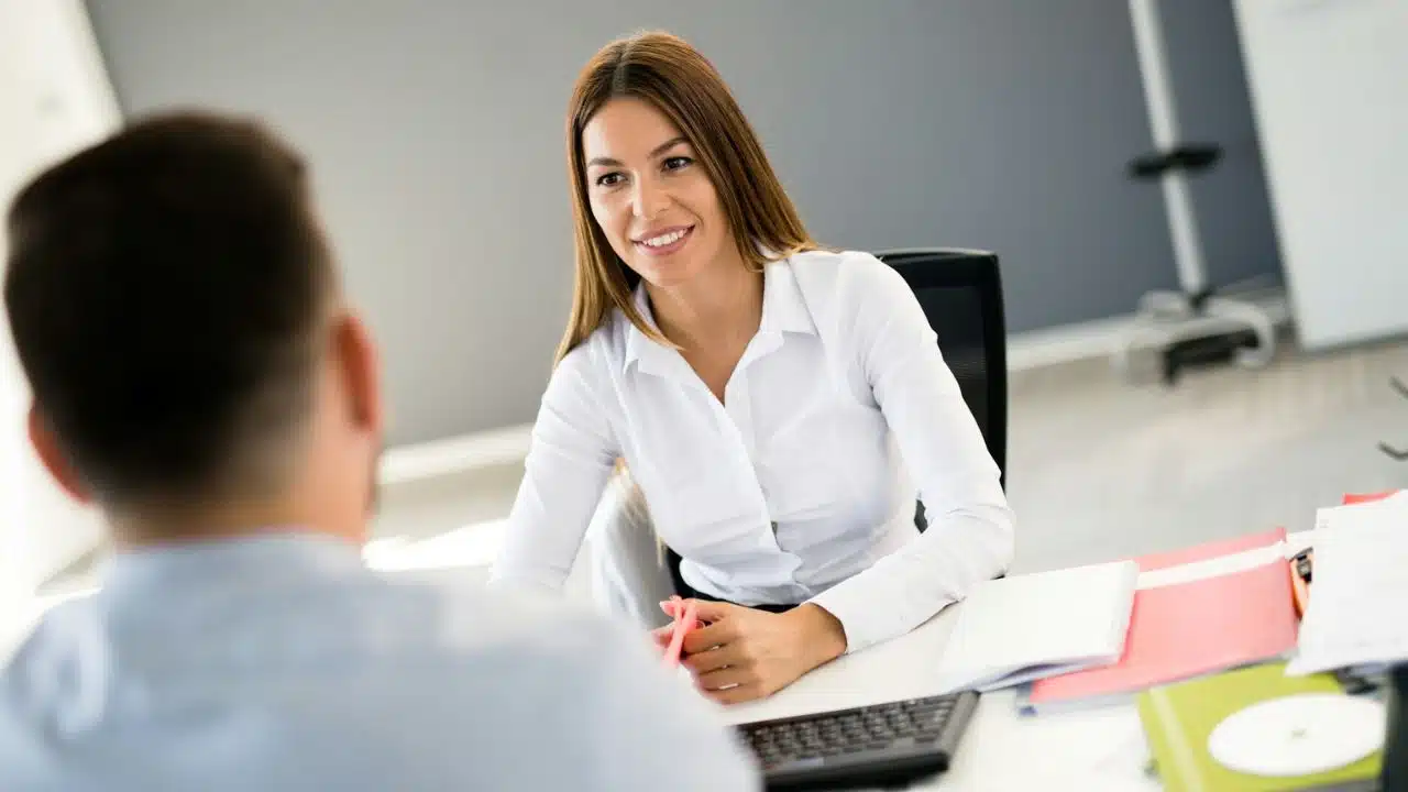 Smiling confident woman in a white shirt sitting at a desk opposite a man.