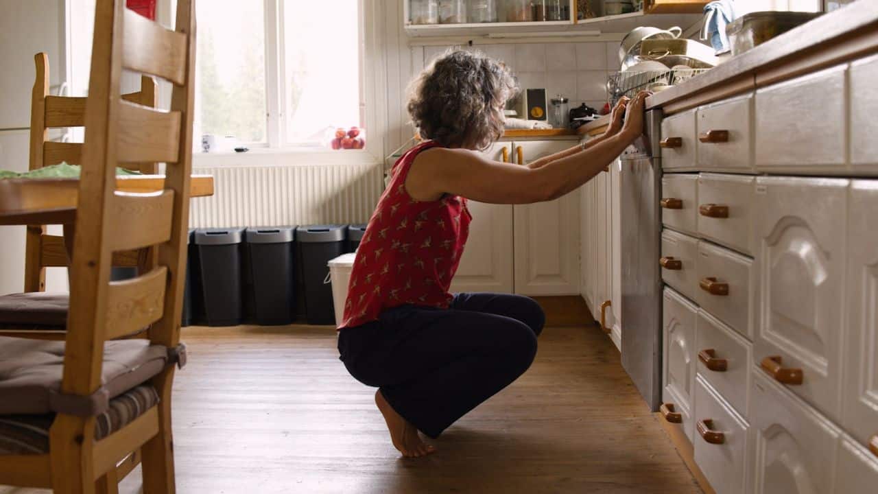 Woman crouching barefoot in a kitchen, looking into a low cabinet or drawer.