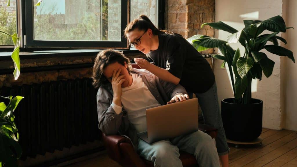 Woman leans over a man covering his face with his hand while working on a laptop.