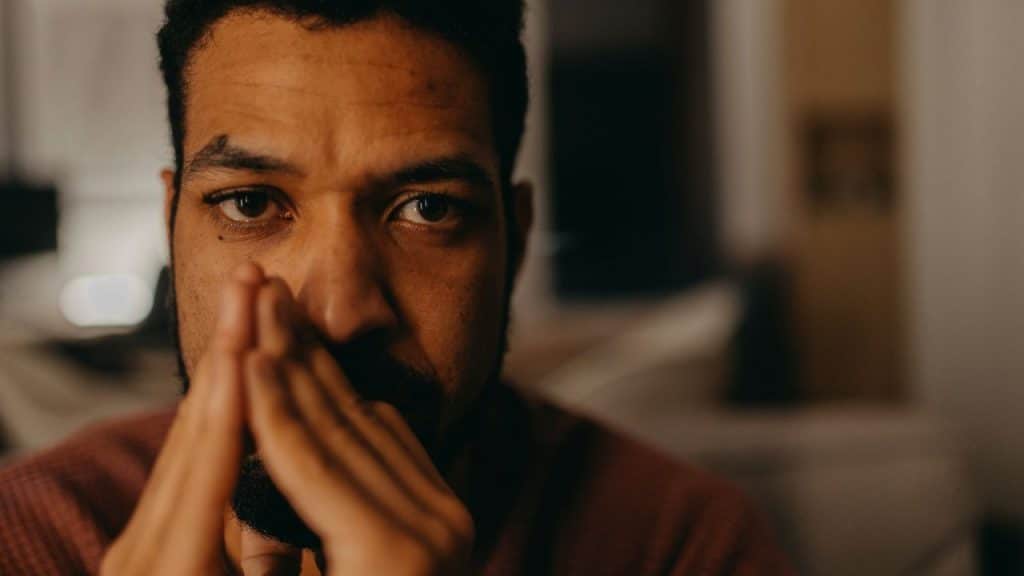 Close-up of a serious man with a beard and furrowed brow, hands clasped near his mouth.
