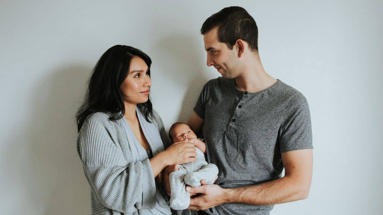 Parents in gray attire lovingly hold and look at their newborn baby against a simple white wall.