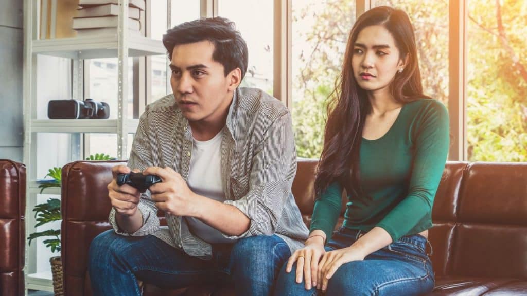 A man plays a video game intently while a woman sits next to him looking bored.