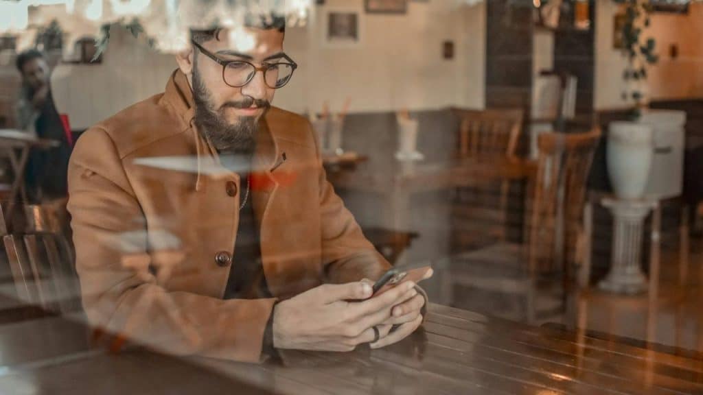 A man in a brown coat sits at a café table looking at his phone through a window.