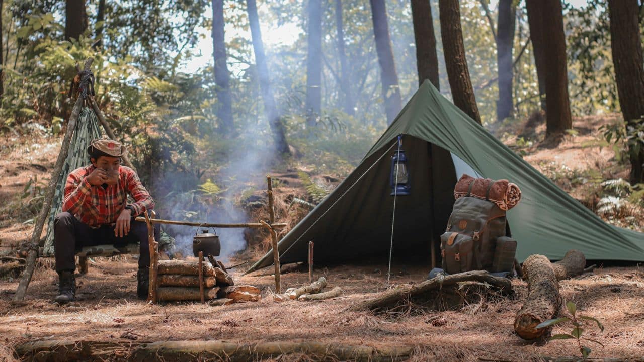 A man in a plaid shirt sits by a camp fire, brewing coffee in a kettle next to a tent in a forest clearing.©Vitaly Gariev/Unsplash.com