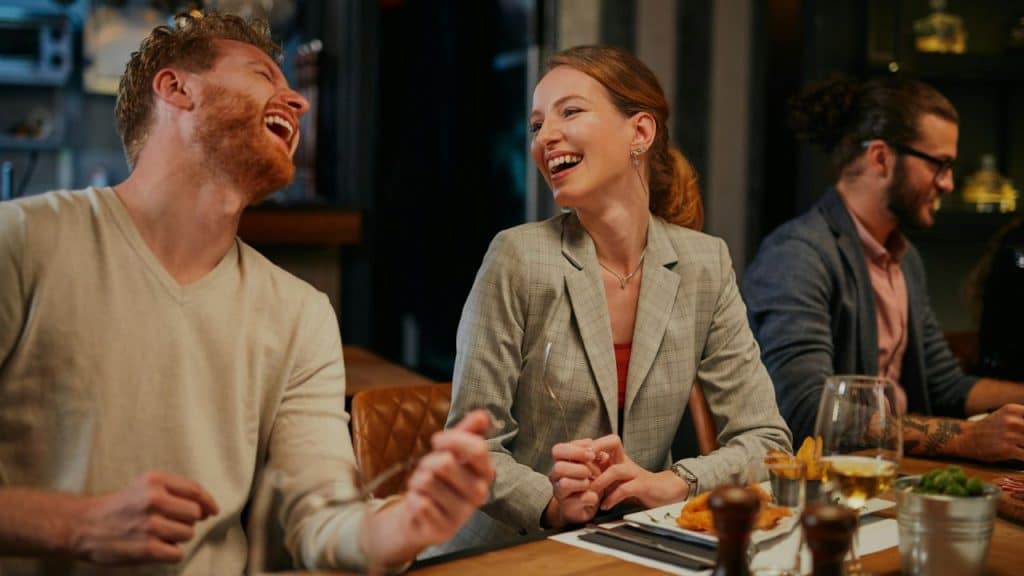 A red-headed man and woman in a plaid jacket laughing together at a dinner table.