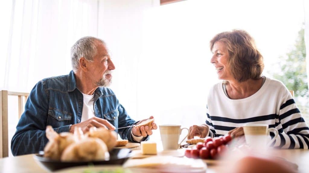An older couple smiles and eats breakfast at a brightly lit dining table.
