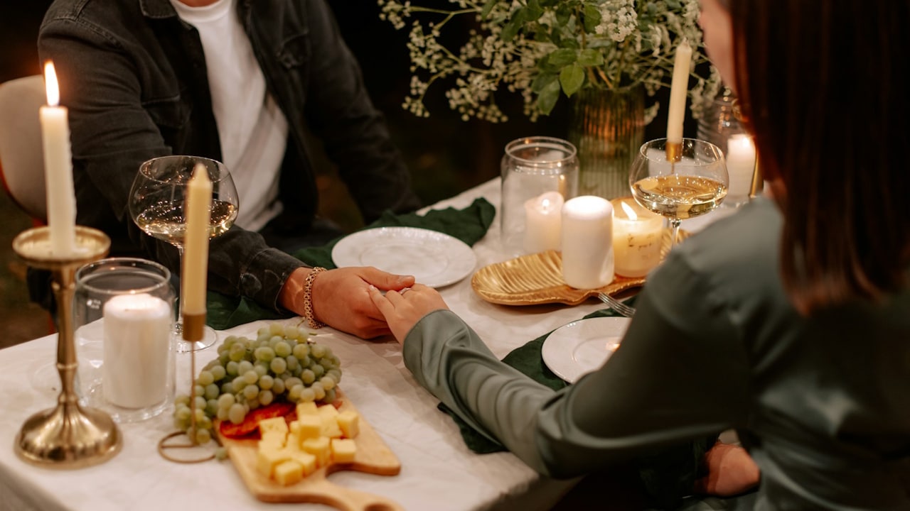 A couple holds hands across a table set with candles, wine glasses, cheese, and grapes during a romantic outdoor meal.