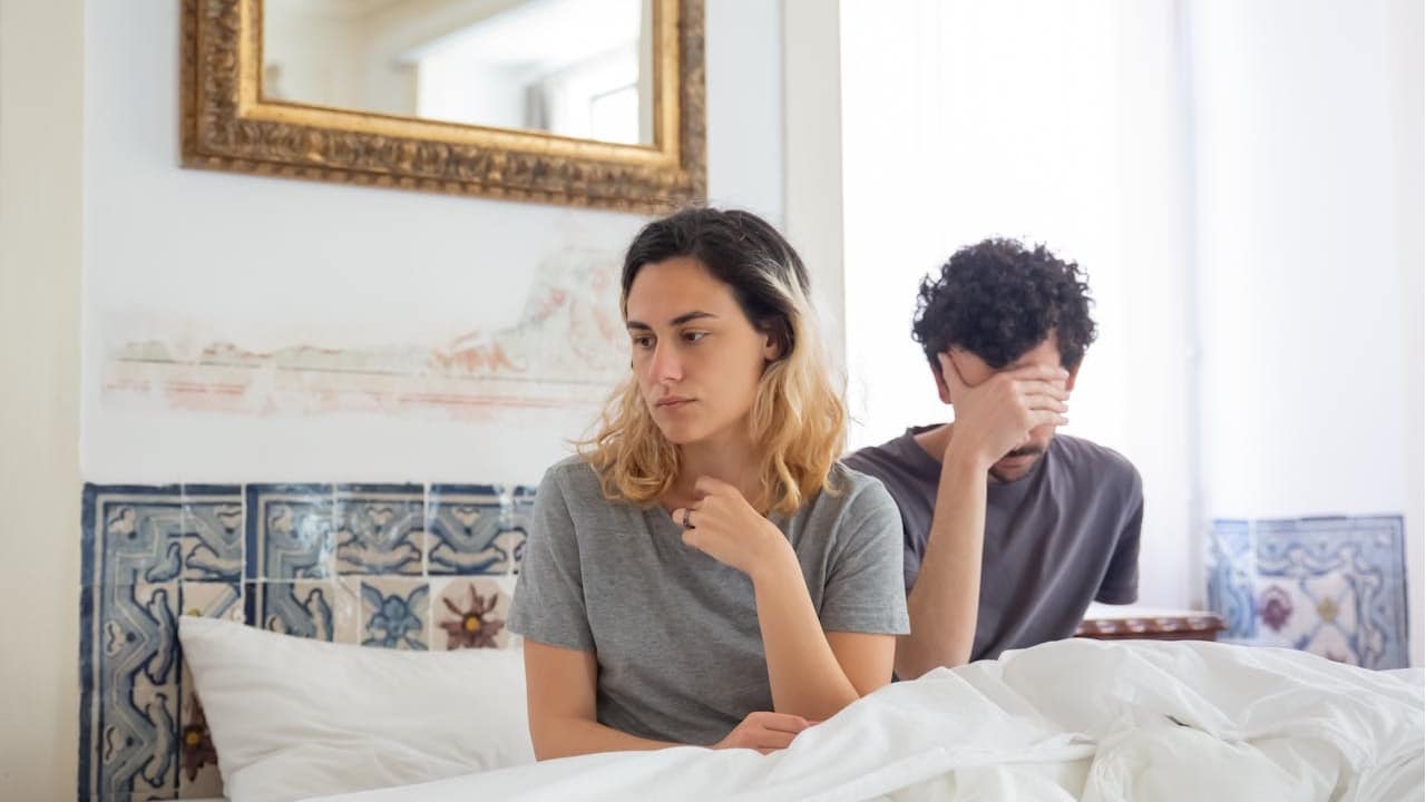 A woman is sitting in bed looking upset while a man behind her covers his face with his hand.
