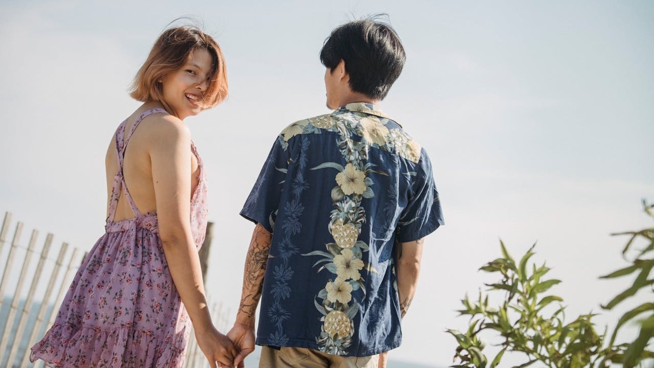 A woman in a floral dress is holding hands with a man in a Hawaiian shirt, smiling back at the camera outdoors.