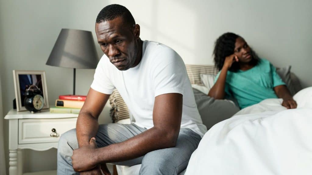 Man sitting on a bed looking down, with a woman sitting behind him.