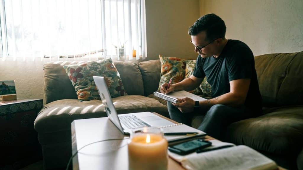 Man working on a laptop and writing in a notebook on a coffee table.