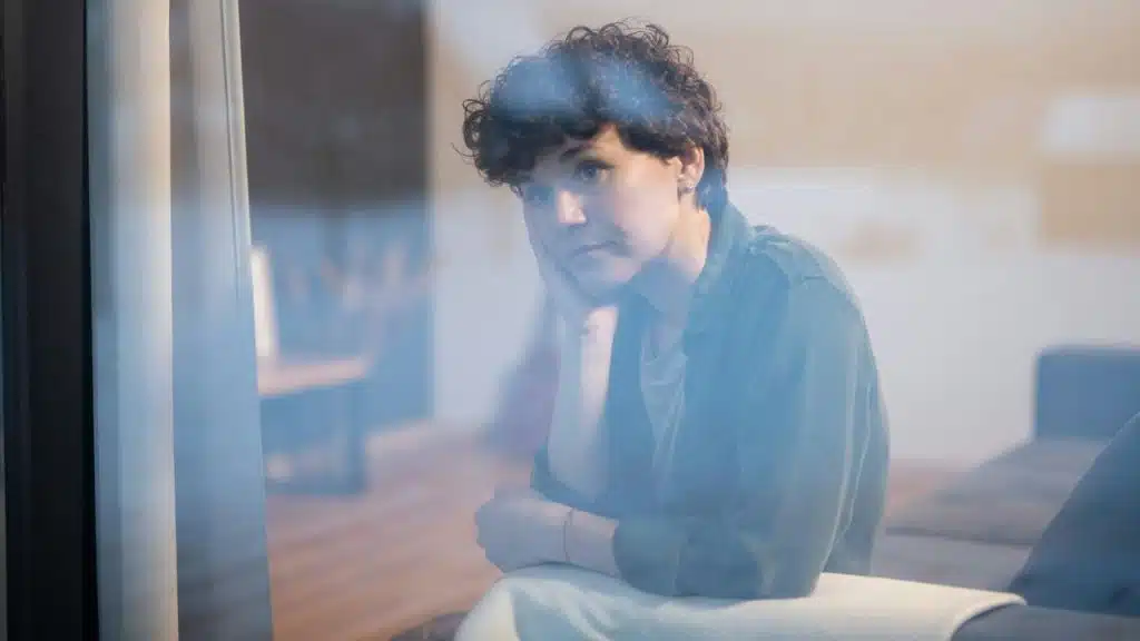 Woman with curly hair looks thoughtfully out a window, resting her head on her hand.