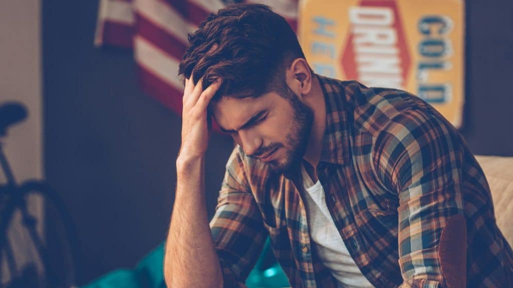 A man in a plaid shirt sits with his head in his hand, looking down.