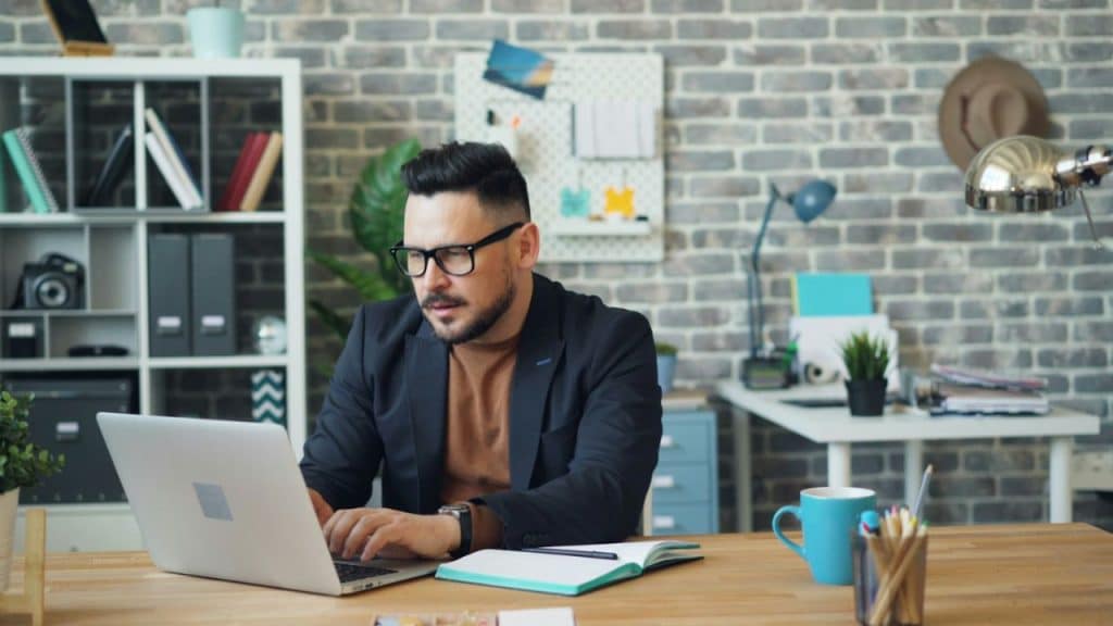 A man in an office works at a laptop with a brick wall background.