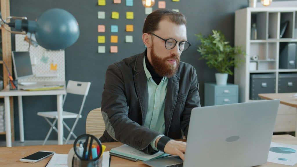 A bearded man in a blazer is working on a laptop in a brightly lit modern office.