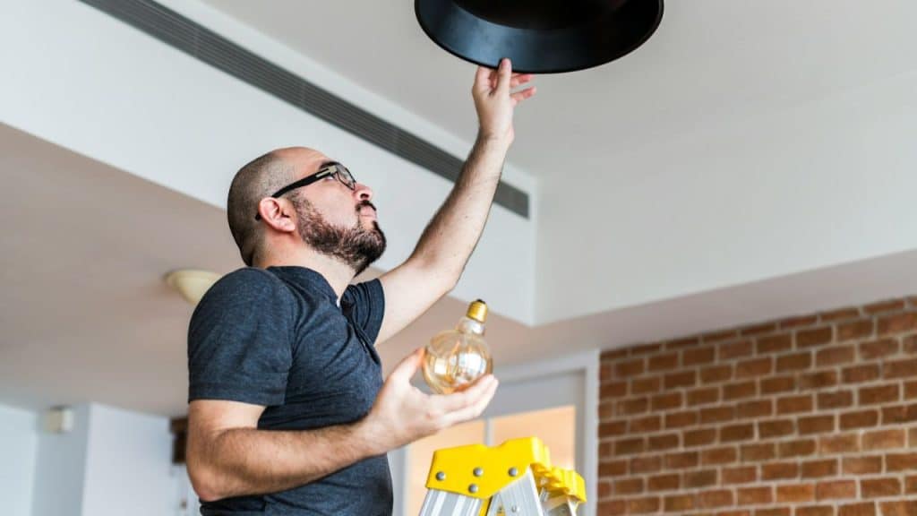 Bald man on a yellow ladder changing a lightbulb in a black fixture.