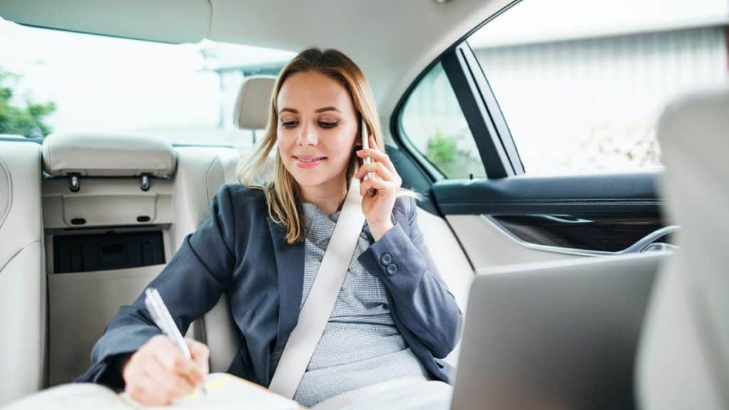 A woman sitting in the back seat of a car talking on the phone while writing in a notebook.