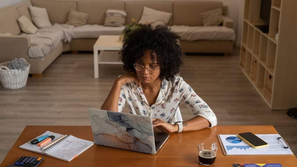 Woman working on a laptop at a table with papers and a drink in a bright living room.