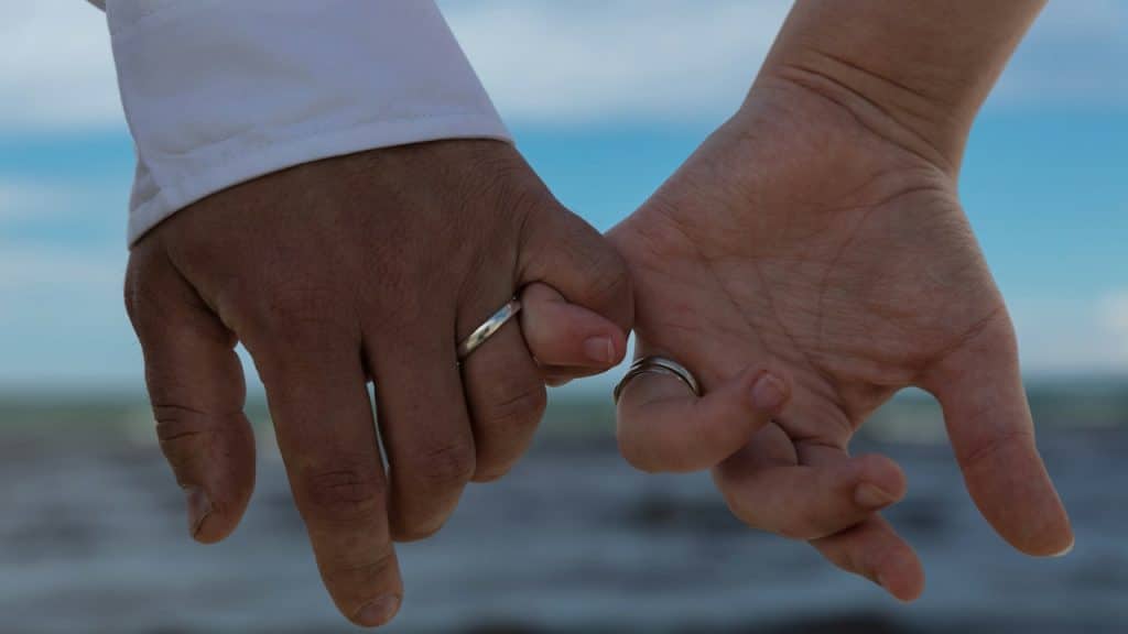 Close-up of two people, one with darker skin and one with lighter skin, holding pinkies and wearing wedding rings, against a blurred ocean background.