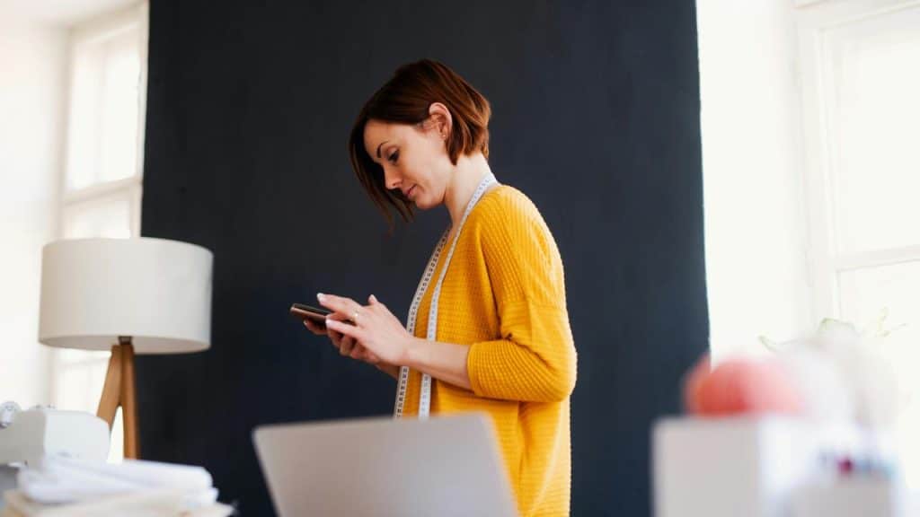 A woman in a yellow sweater with a measuring tape around her neck looking at her phone.