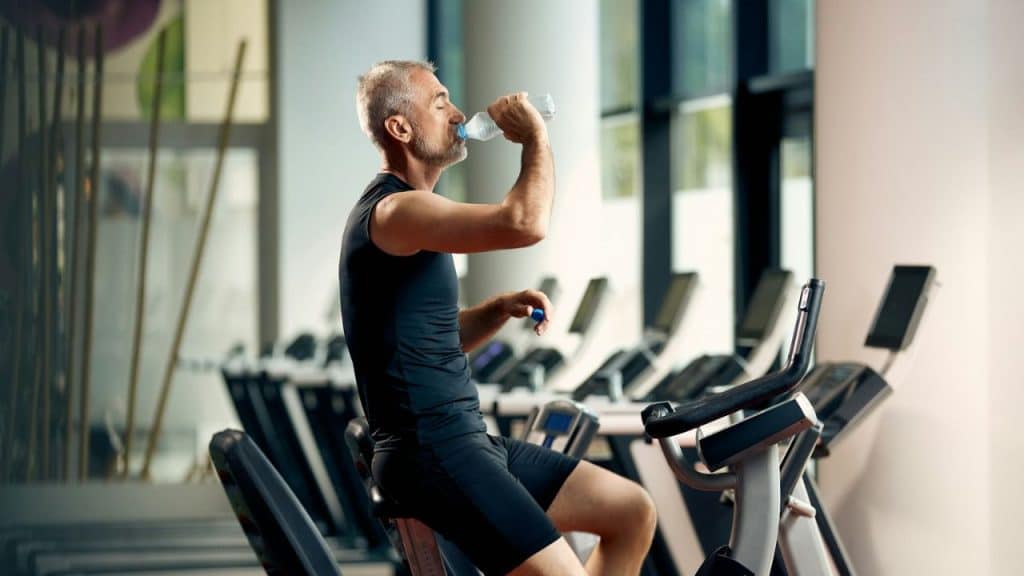 A middle-aged man on an exercise bike drinks water inside a gym with treadmills.