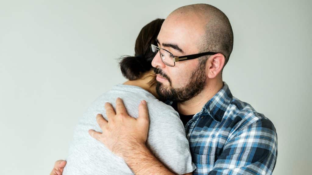 A man with a beard and glasses embraces a person in a gray shirt against a plain background.