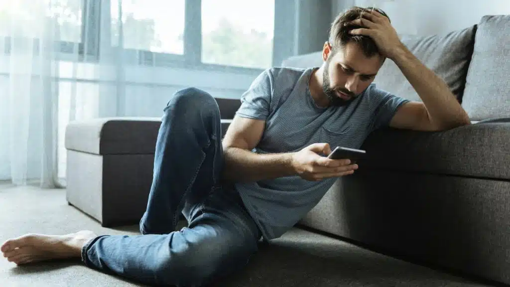 Barefoot man leaning on a couch, looking down at his smartphone with a worried expression.