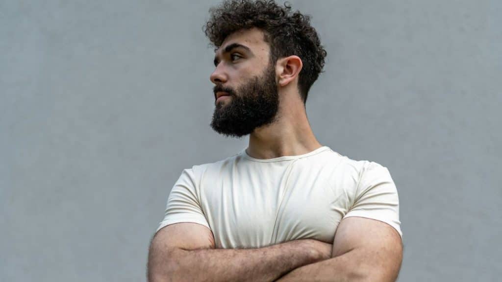 A man with curly hair and a beard stands with crossed arms against a gray background.