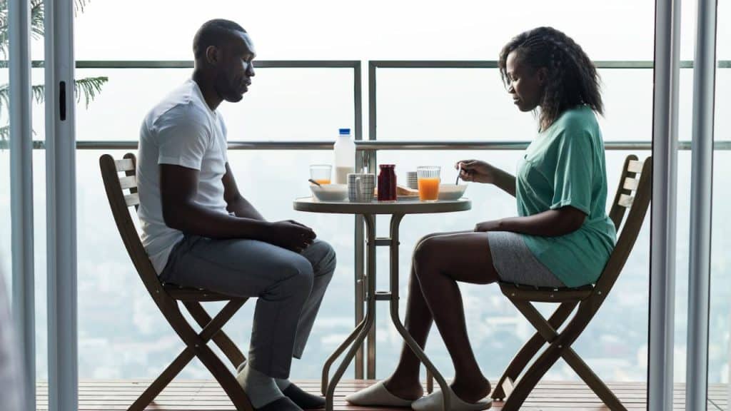 A man and a woman sit silently facing each other at a small table on a balcony.