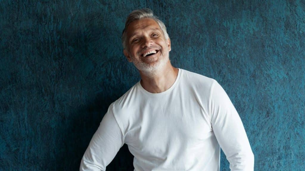 Man in a white long-sleeve shirt smiling broadly against a blue textured wall.